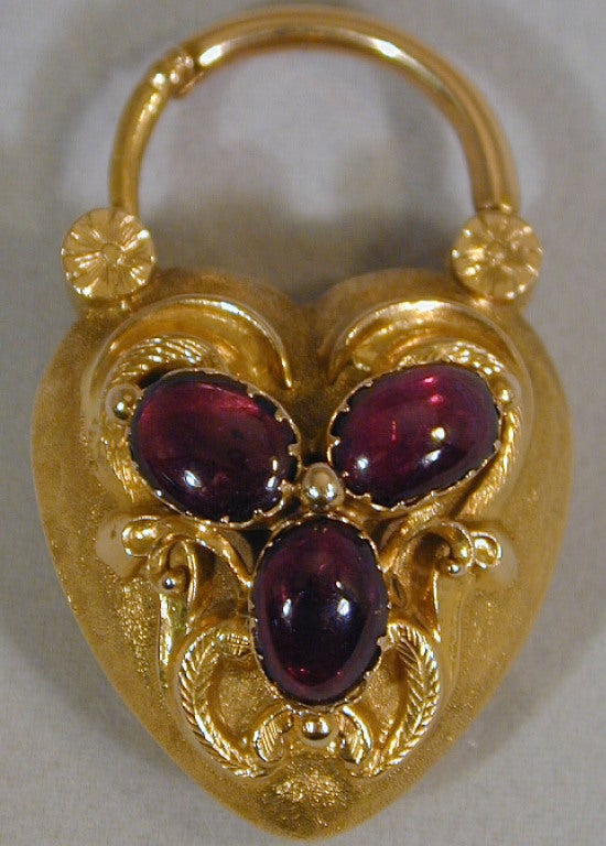 Elaborately carved 18K heart lock set with three cabochon garnets can be worn as a locket or used to hold a linked bracelet. The compartment on the reverse holds a lock of woven hair, a memento of a loved one.