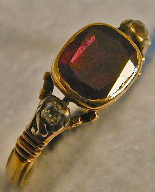 Georgian almandine garnet ring in an 18K gold basket setting flanked by two cushion cut diamonds. Almandine garnets are purply red and were flat cut as seen here. This lovely early ring is a size 6 1/4 and the stone measures 5/16