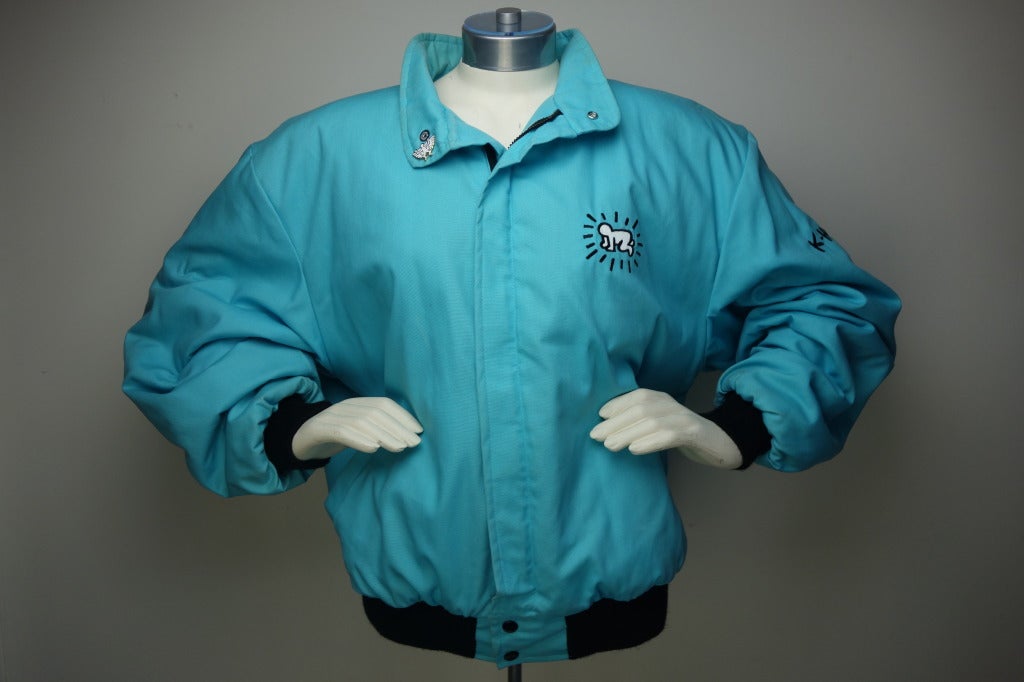 Keith Haring designed by Identity Inc. NY. turquoise jacket, two front pockets, red quilted interior with one interior pocket and a Keith Haring white enameled pin.