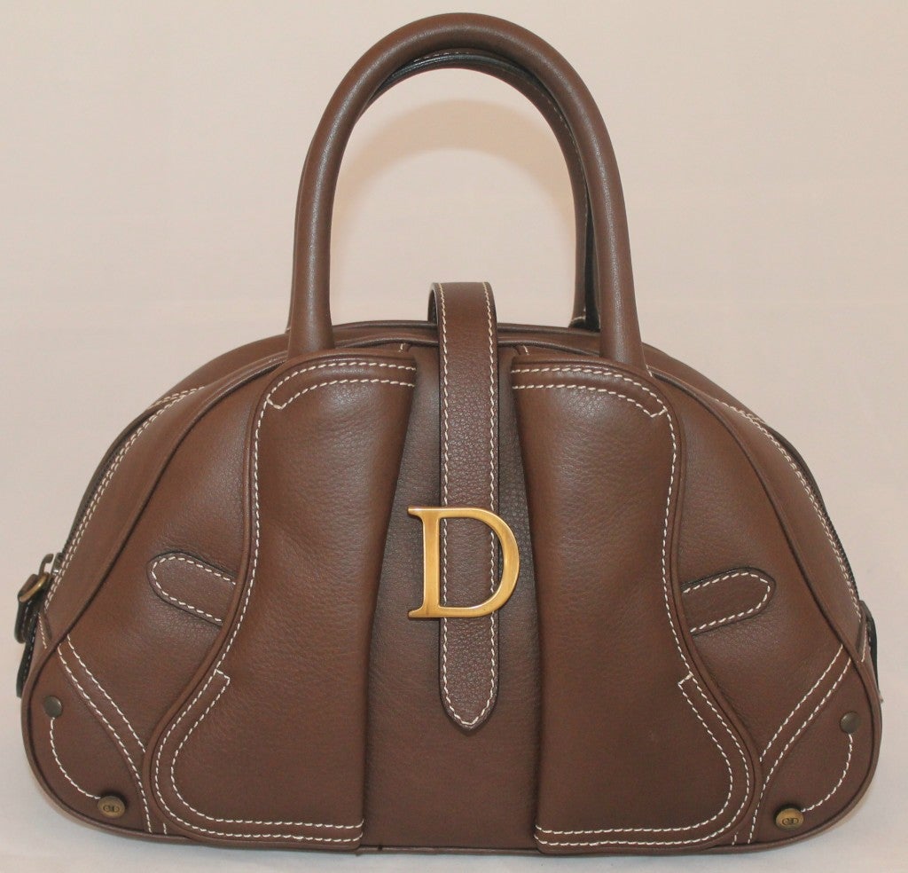 This Christian Dior brown leather mini bag is structured with a top handle. It has gold hardware, white stitching, one exterior compartment, and one zip interior compartment. There is a zipper closure with a flap closure on top that has the 