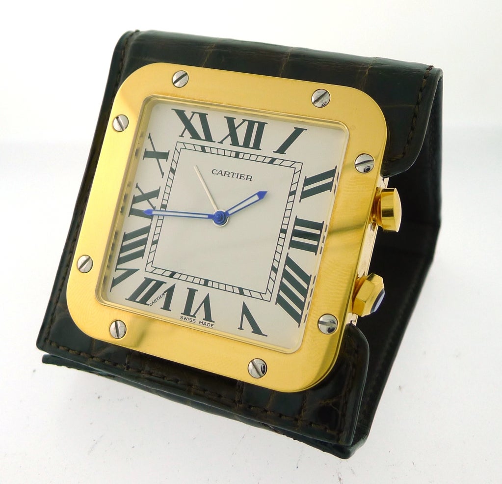 This is the Cartier Santos Travel Alarm Clock. It has a Brown Croc Skin cover and a quartz movement. The case material is Yellow Gold and Stainless Steel. The size is 77 x 65mm closed.