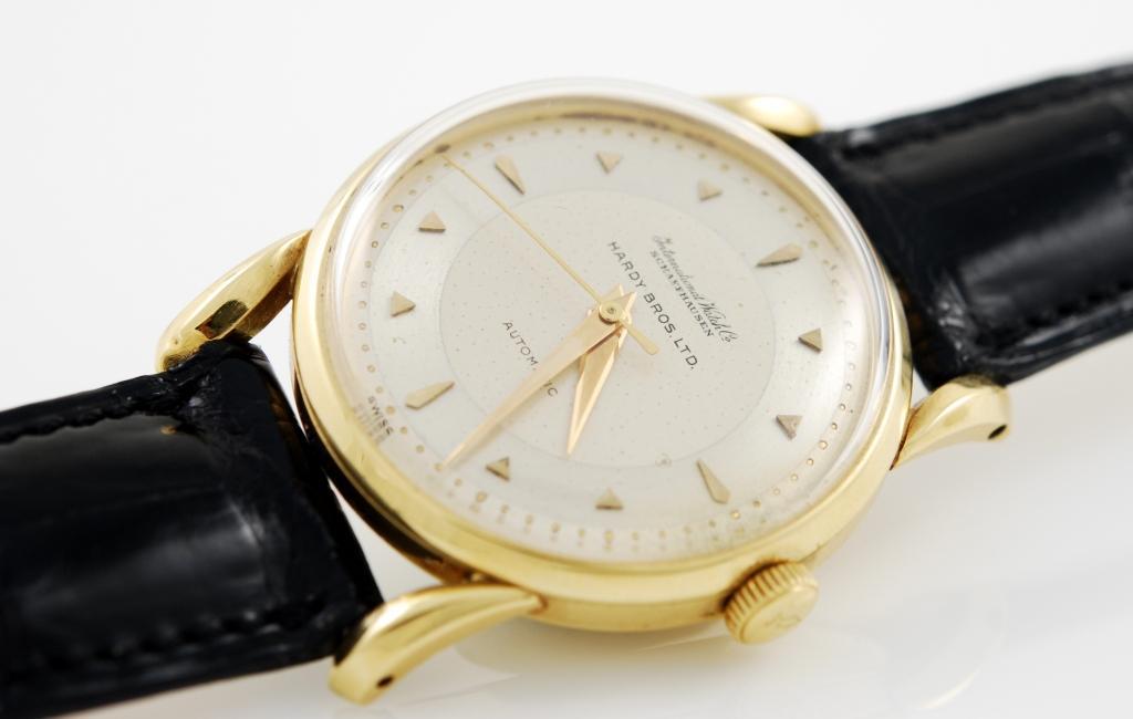 18k yellow gold IWC wristwatch, circa 1950s. Two-tone dial, signed Hardy Bros Ltd., with gold applied faceted indexes and dauphine hands. 35mm diameter case with plastic crystal, automatic movement, caliber C852. Leather strap.