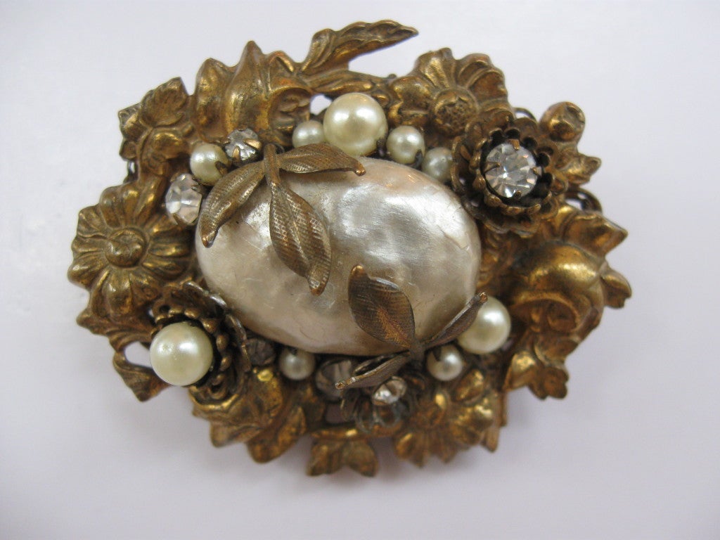 This fine example of Miriam Haskell's design and craftsmanship, features an oval brooch with a large center baroque pearl, surrounded by gold metal, smaller pearls, and paste.