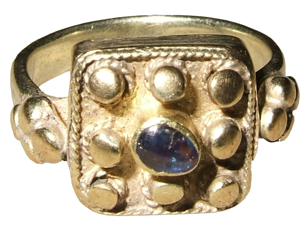 electrum and sapphire
Bezel 12 x 12 x 7 mm.; circumference 58 mm.; weight 7.4 gr.; US size 8 ½; UK size Q ½ 

The basic form of the ring, the clusters of globules at the shoulders, and the workmanship of the bezel, where the globules of the