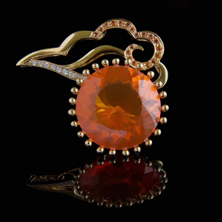 An 18K gold pendant set with a 24.91ct Brazilian fire opal and featuring an Asian cloud motif design inlaid with orange sapphires (0.50ct) and diamonds. A pin on the reverse allows it to also be worn as a brooch/pin. The detachable, variable length