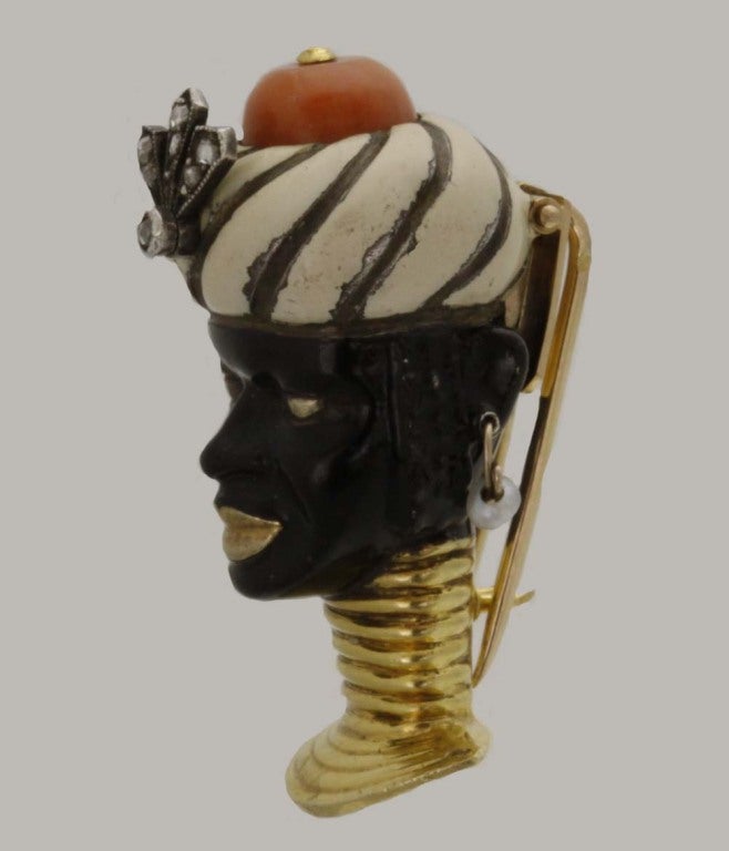 This Art Deco brooch features a black enamel face with gold features, and wears a black and white turban topped with diamond-accented coral. With gold ribbed neckwear. Numbered 0557.