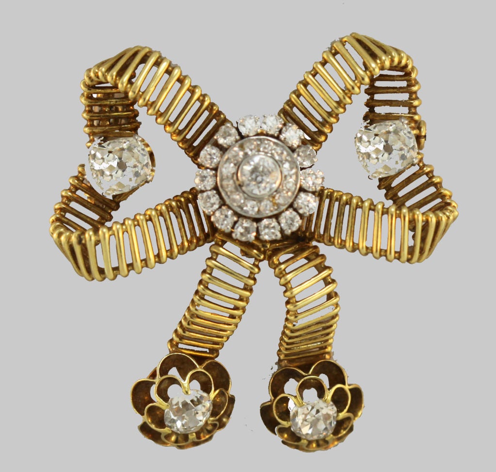 Mellerio latticework forms a stylized bow brooch , with a centered round-cut diamond surrounded by two rows of smaller round-cut stones. Each curved bow side features a larger cushion cut diamond, while the ribbon “tails” end with a cushion cut