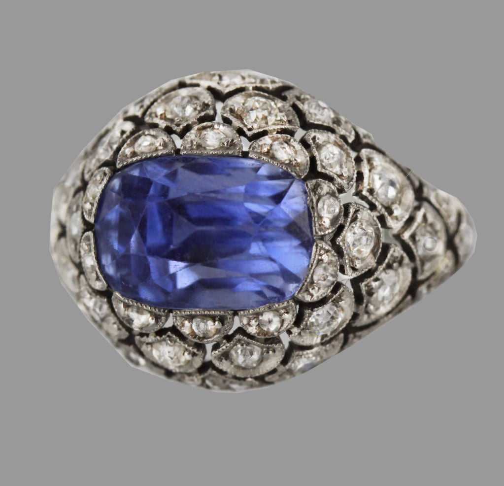 This unusual Edwardian ring features a stunning Ceylon Sapphire of almost 6 carats in a ring comprised of round-cut diamonds set into an allover scallop-patterned platinum setting. Top of shank features delicate open scroll filigree.
