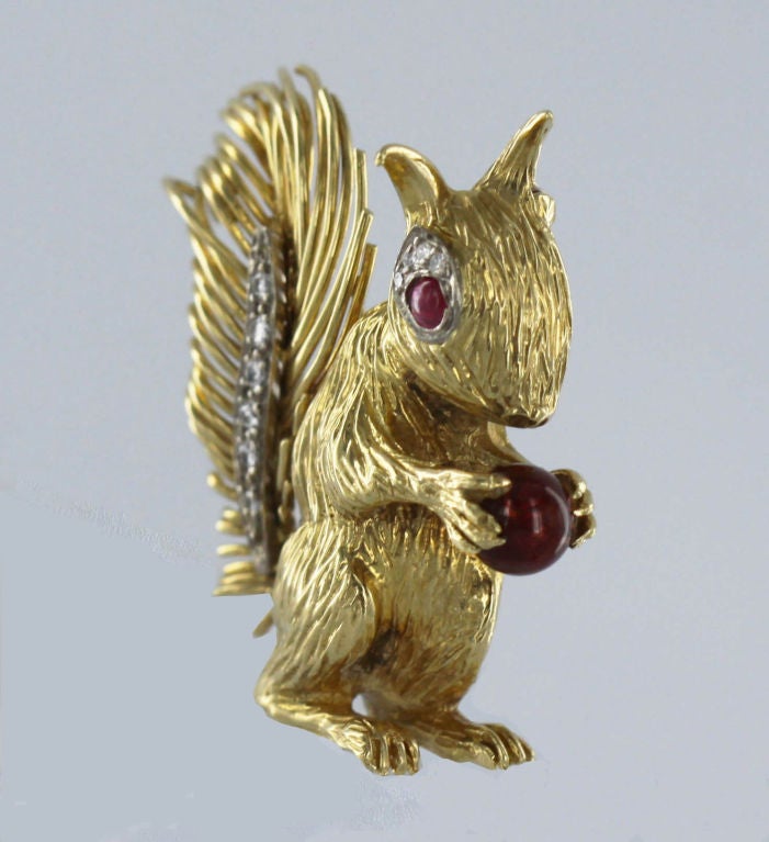 This charming 18K gold squirrel designed in 1967 by the English firm Kutchinsky sits up in silhouette, holding a ruby bead in delicate paws while looking out from a ruby cabochon eye edged in diamond details. The tail of swirling gold fur has