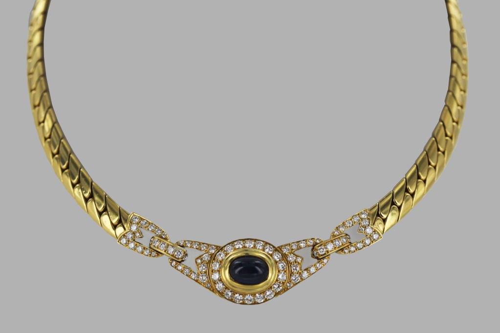 A Cartier necklace from the 1980's. The necklace is an 18K gold herringbone chain which showcases a center cabochon sapphire set in gold, and encircled by a single row of diamonds. On either side of this dramatic blue stone are two geometric,