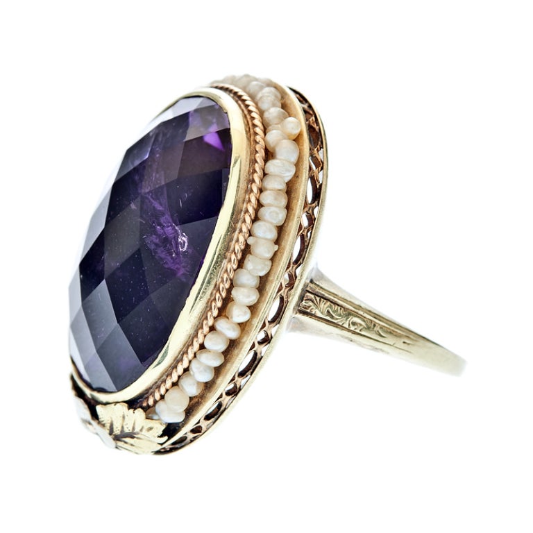 A large, faceted, oval amethyst exhibits deep, luscious color with a surround of tiny, natural seed pearls wired to its frame in this outstanding antique ring. Floral motif and delicate filigree add whimsey to this statement ring