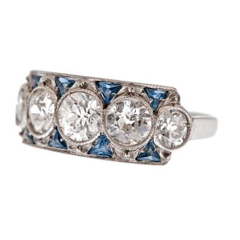 Lovely art deco ring, consisting of five ideally matched old European cut diamonds, 1.80 carats in total, bezel set and accented with eight trillion cut blue sapphires. This ring has an important presence on the finger, however, it lays perfectly