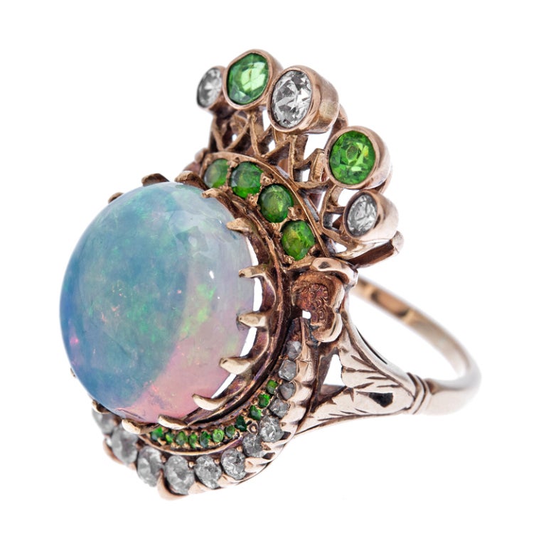 Extremely unique ring, designed to resemble a crown, consisting of a 14mm cabochon opal in a scalloped claw setting, framed by a crescent shape of demantoid garnets and diamonds on one side and bezel set diamonds and demantoid garnets on the other.