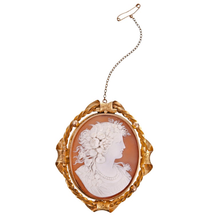 This superb shell cameo depicts a beautiful female form with an expressive gaze and flowing hair adorned with intricately carved flowers. The oval cameo is ornately framed with a textured bezel and curling rope forms that twist and curve. Extremely