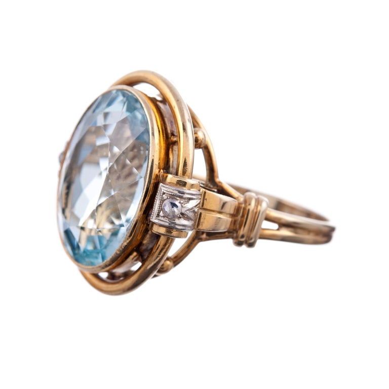 A charming piece of hand fabricated jewelry with a faceted oval aquamarine of 10 carats surrounded by a double bezel of gold and accented with two modest diamonds. The mounting has an ornate scrolling design and allows the major stone to sit up and