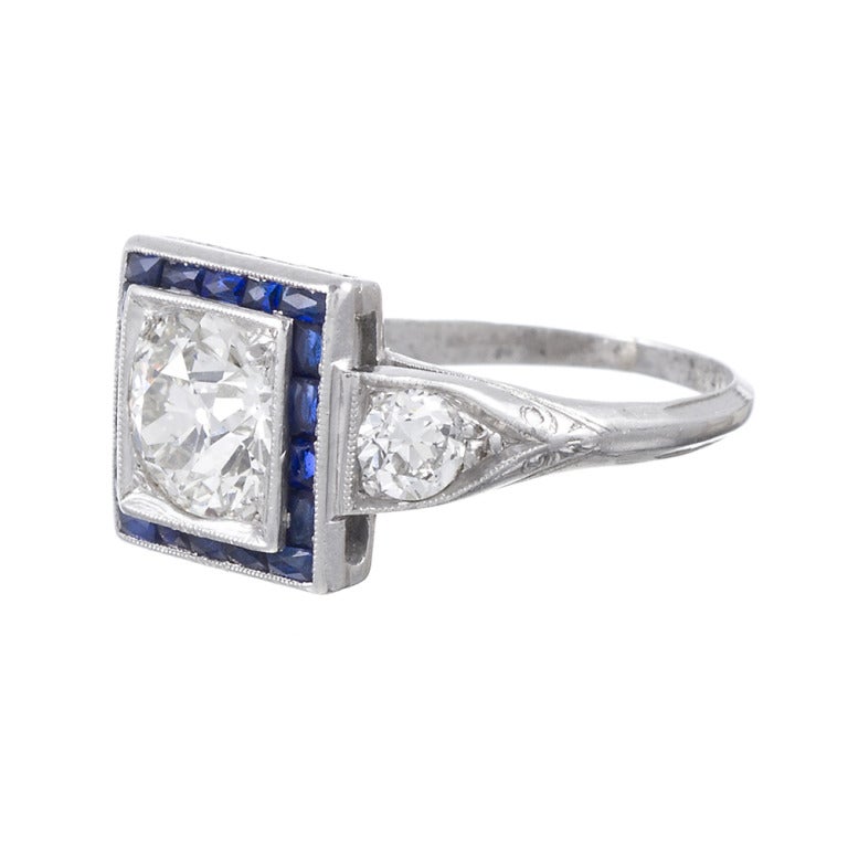 Platinum art deco diamond ring, celebrating the distinctive details that deco enthusiasts celebrate: striking contrast between white diamond and blue sapphire, architectural style elements and abundant detail. The center diamond is old European cut,