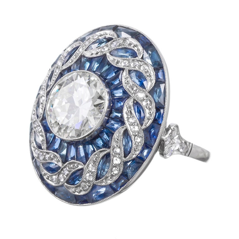 Platinum ring, set in the center with a 2.70 carat round brilliant diamond. We grade the diamond as J-K color and Vvs clarity. The center stone is surrounded by a twisted diamond frame and custom cut blue sapphires, forming a three dimensional