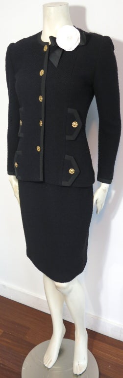 Vintage ADOLFO 1970's Classic black knit skirt suit with engraved gold buttons, grosgrain ribbon trims throughout, and left neck camellia bow tie embellishment.  Double pocket flap detailing at both sides.

Elasticated waistband in matching