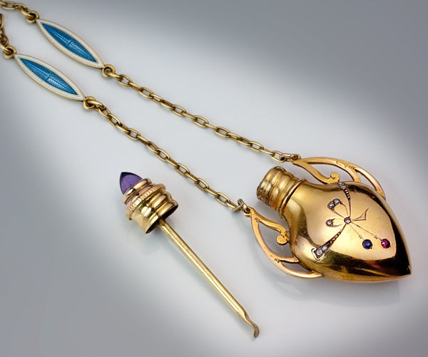 The perfume flask is shaped as an amphora with Art Nouveau handles and gem-set engraved decorations.  The cap is set with a cabochon amethyst. 

The gold chain with six double-sided inserts of blue guilloche and white opaque enamel.

Made by