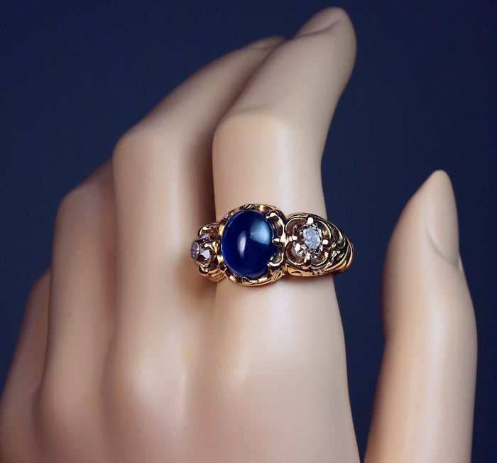 This Renaissance revival antique three stone gold men’s ring was made in St. Petersburg between 1899 and 1904.

The ring is cast in high relief with renaissance style scrolling foliage and prong set with a large blue cabochon sapphire
flanked by two