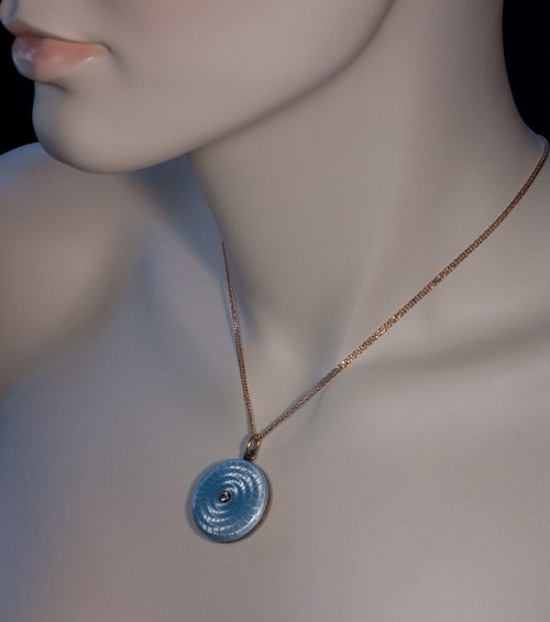 The locket is crafted in gilded silver and gold and completely covered with delicate pale blue guilloche enamel. Wavy lines radiate from the center, simultaneously creating a sunburst pattern and a spiral effect. 

The cover is centered with an