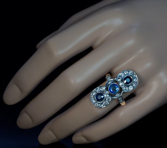 The ring is vertically set with 3 natural blue sapphires surrounded by 26 old mine cut diamonds and 4 rose cut diamonds.
The gemstones are set in silver over 14K gold. 
The central medium blue sapphire is approximately 0.77 ct, the pair of dark