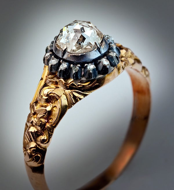 The ring is centered with a foil-backed old rose cut diamond (5.9 - 5.5 x 3 mm, approximately 0.80 ct) in a silver cut-down closed back setting,  surrounded by a row of tiny rose cut diamonds set in silver.  The shoulders of the ring are elaborately