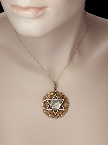 An original pre World War II Art Deco pendant designed as a round openwork matte gold plaque with a white enamel six pointed Star of David set with a hexagonal aquamarine and six small rubies. The star is flanked by six Art Deco openwork floral