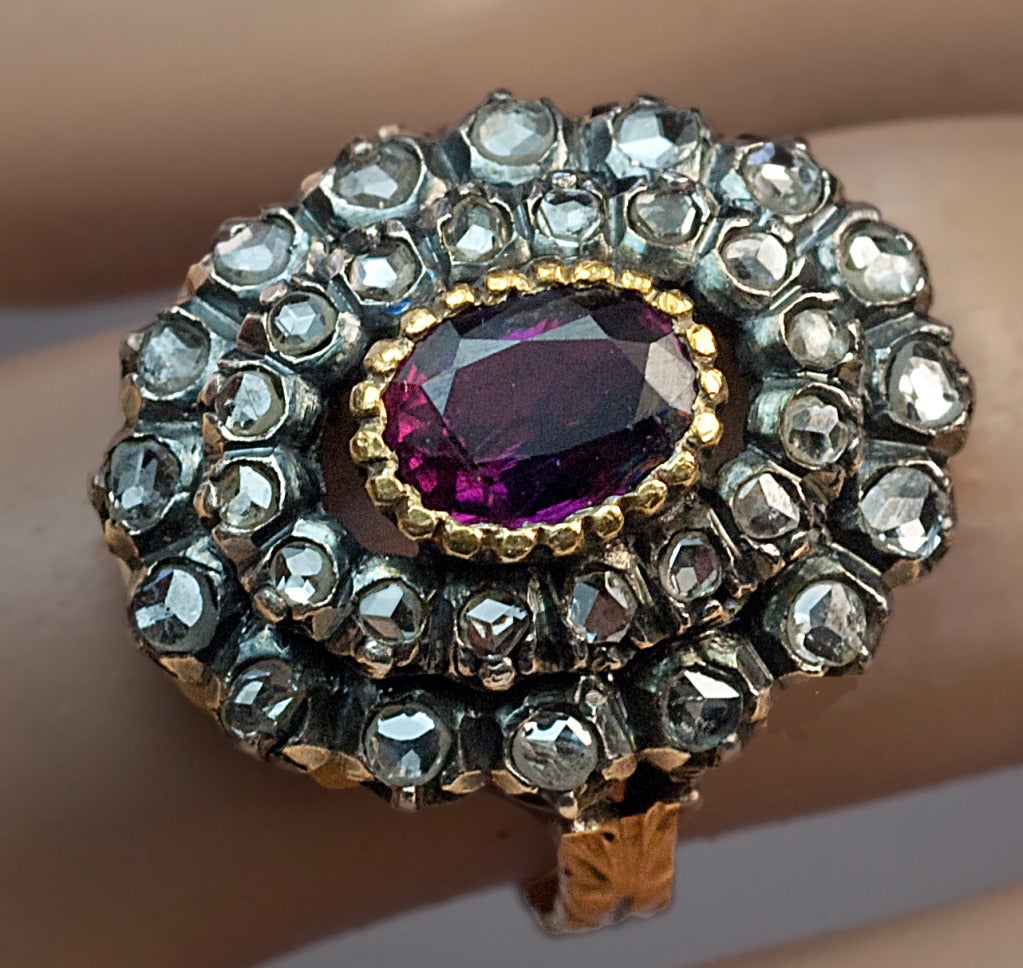 An Antique Georgian Era Ruby and Diamond Cluster Ring

circa 1800

The ring is centered with an oval ruby of a deep reddish purple color (7 x 5.2 x 4.1 mm, approximately 1.19 ct) set in a fluted gold topped silver bezel surrounded by a double