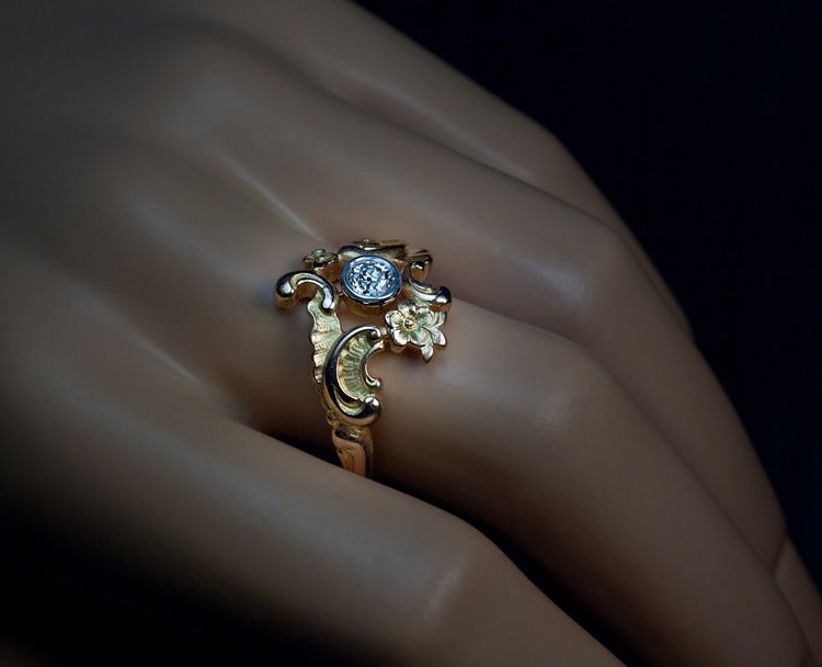 This highly unusual  Russian 14K gold openwork ring is centered with an old European cut diamond (approximately 0.32 ct) set in a silver and gold bezel held by two rococo chased two-tone scrolls embellished with flowers in Art Nouveau taste.

Made