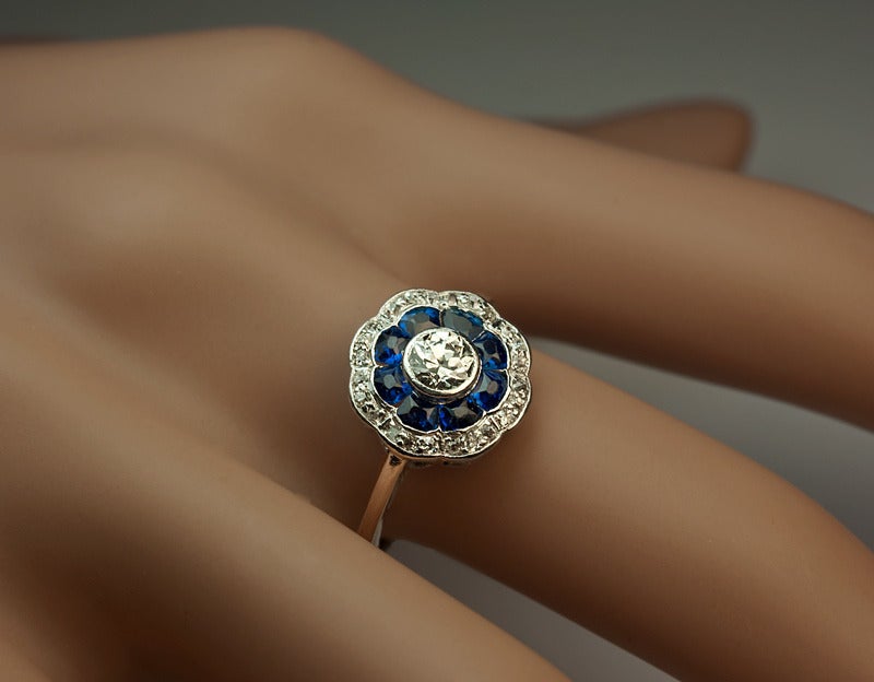 circa 1910

The ring is centered with an old European cut diamond (approximately 0.35 ct) surrounded by a row of tapered calibre cut natural sapphires (approximately 0.50 ct tw) and further set with a border of old cushion cut diamonds (estimated