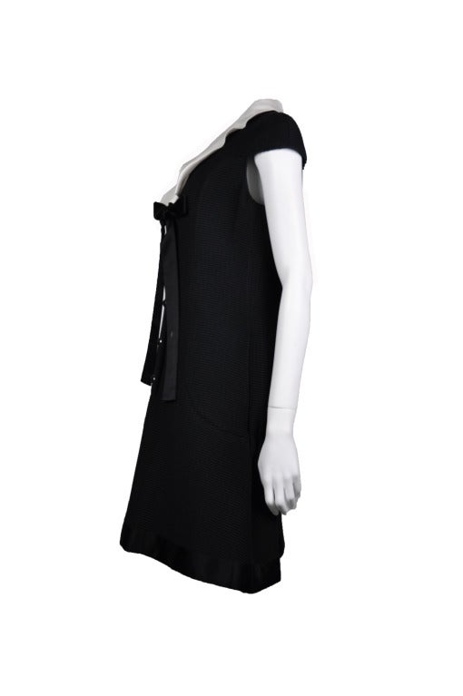 This knee length Chanel black dress has a white silk fold down collar with a black silk ribbon neck tie.  Pewter tone CC logo button fastening at front.  Two pockets on sides. Fully lined in silk. Made in France.
Material: 81% cotton, 19% nylon