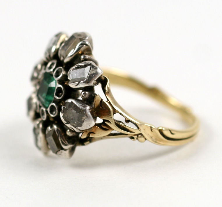 A cushion cut emerald sits at center of this 15kt gold ring. The emerald is surrounded by old cut diamonds of approximate .80cts in weight in a flower head form. All stones have been foiled and set in closed rubbed over settings. The shank continues