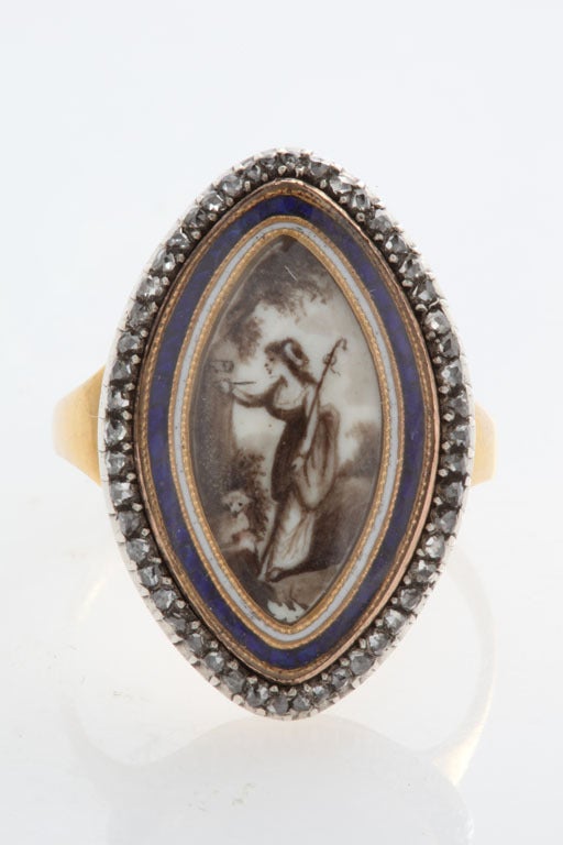The jewel is a navette shaped lover's ring with sparkling rose diamonds on the outer perimeter. Gold frames both blue and white inner enamel borders. Under pure rock crystal, a youthful shepherdess, smiling in thought,  is intent upon carving the