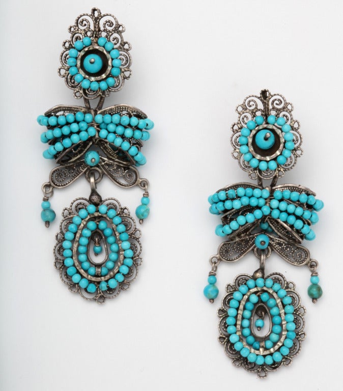 These spirited chandelier earrings are typical of Colonial Mexican design. The silver is worked in curved, substantial silver wire. The spiral silver swirls are known as cannetille work. A flower at top suspends a layered bow that is tasseled with