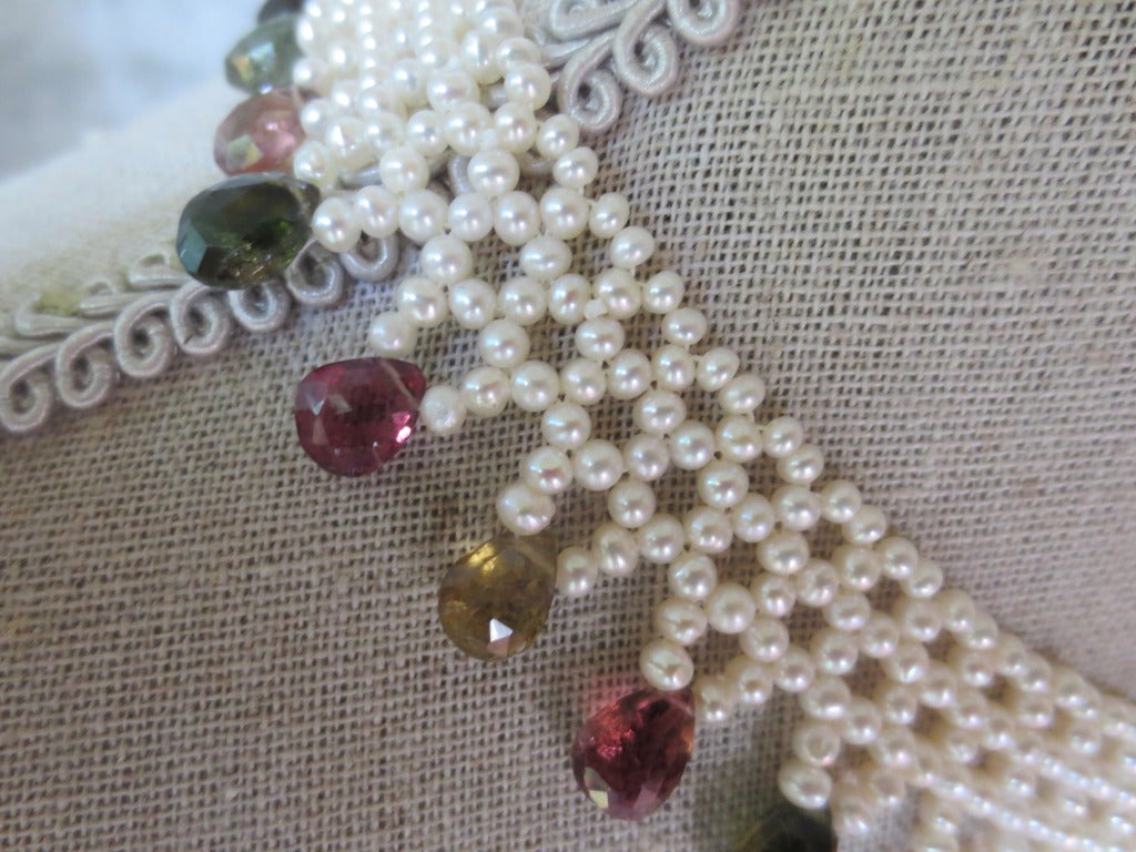 This hand woven necklace is made using 2 mm cultured pearl beads to create a lace-like design that perfectly fits the collar. Necklace is tapered in design to fit along the curve of the neckline. Graduated multi colored tourmaline briolettes hang