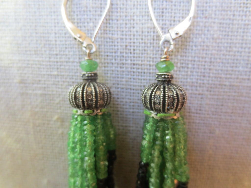 These onyx and tsavorite tassel earrings with diamonds and 14k white gold lever backs are striking and elegant. The tassels hang from a diamond-encrusted sterling silver crown-shaped cup. Both lever backs are made of 14k white gold, highlighting the