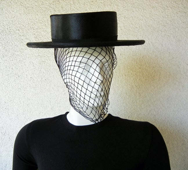 Adrian Original black Cordobes hat designed with the fashion flair of the Spanish style equestrian hat.  Black net attached at front drapes over face.  While the hat has the look and sheen of black satin it could also very well be fashioned of