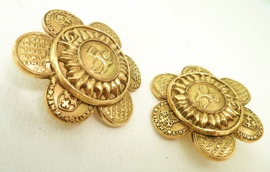 These vintage signed Chanel  “sunflower” earrings feature a centerpiece surrounded by circular medallions bearing the CC shield and address in a gold-tone setting.  In excellent condition, these clip earrings measure 1 ½ inches in diameter and are