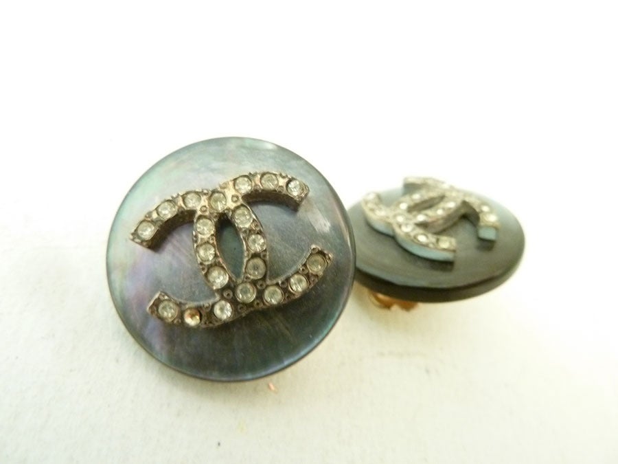 These vintage signed Chanel 96A earrings feature the famous CC logo with clear rhinestones on a pearled-gray resin backing in a silver-tone setting.  In excellent condition, these clip earrings measure 7/8” in diameter and are signed Chanel 96A.
