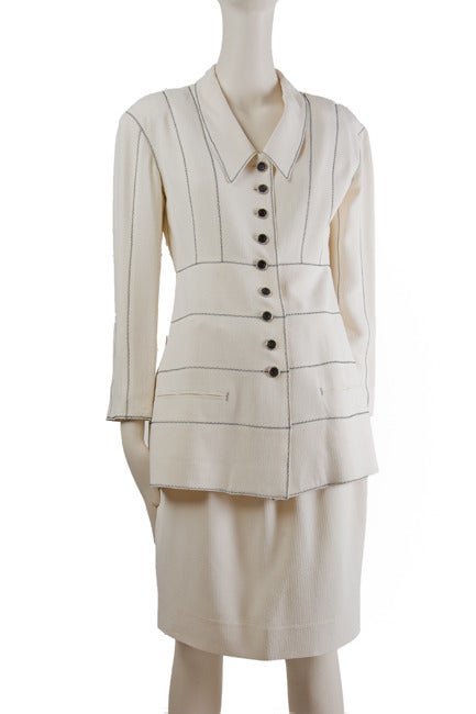 Jacket in creme viscose/acetate by Karl Lagerfeld for Henri Bendel features princess top seaming in a black zig-zag stitch, v-neckline, elongated chelsea collar, long sleeves, two lower welt pockets, and nine button front closure. Skirt features