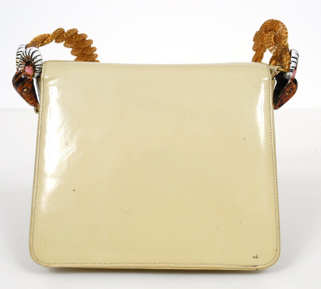 Handbag presented by Judith Leiber in a cream patent leather with gold tone hardware and clasp. This handbag features a unique strap made of braided gold chain and two ram heads connecting the strap to the handbag. The ram heads are said to be