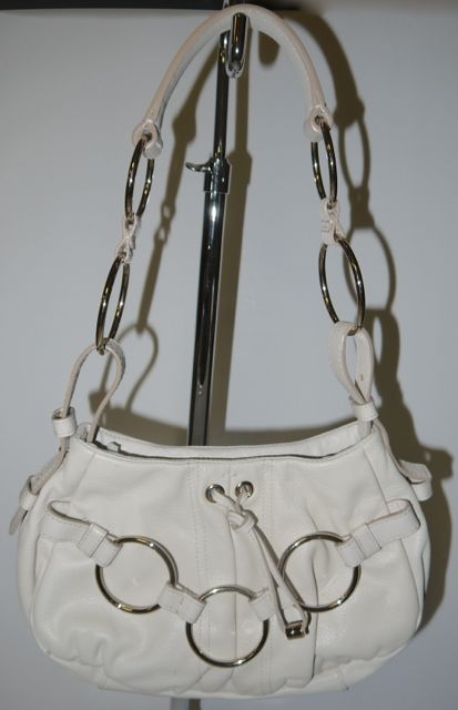 This bag is presented in off white leather and features silver tone hardware.  It has a large, lined compartment with a separate zipper compartment inside and snap closure. Large silver tone rings and leather buckle straps comprise the handle.  This