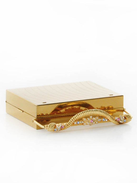 Minaudière compact by Volupte features gold tone case, 4 separate interior compartments include lipstick, powder puff, tissue, flip mirror; and decorative floral rhinestone hinged clasp closure. Black faille frame features. There is a silk