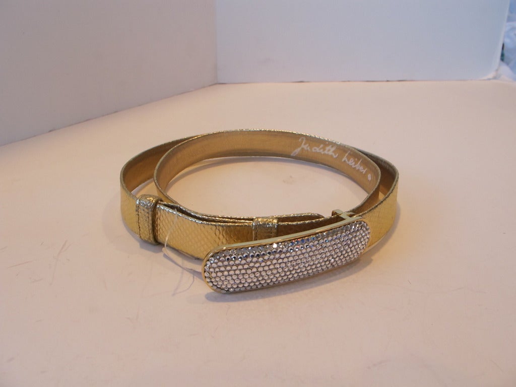 Buttery soft gold leather belt in lizard design. Belt is adjustable and accented by Iconic Judith Lieber  rhinestone buckle which measures 3.5 inches long by 1 inch wide. 1 inch wide belt. The belt measures 43.25 inches total length. The belt is