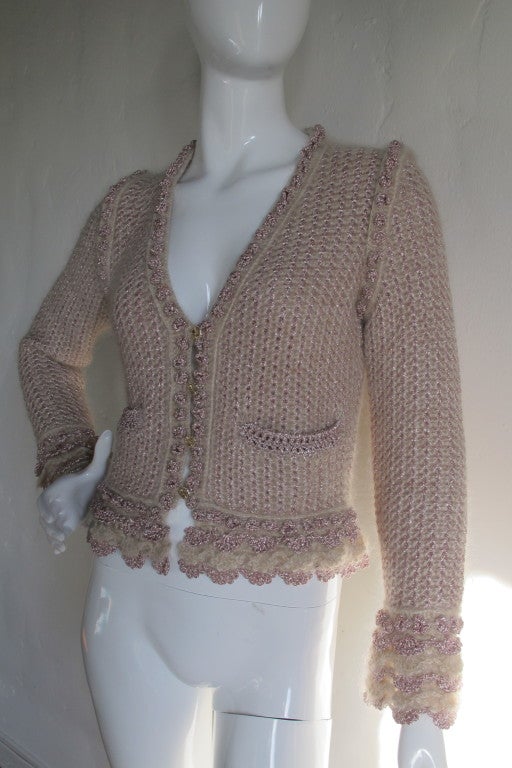 2004A Chanel mohair blend cardigan in a oatmeal hue with pink metallic lace crochet detail at sleeves and bottom edges. The cardigan closes by means of four metal buttons down the center and feature a raised image of the sun with the cc logo in the