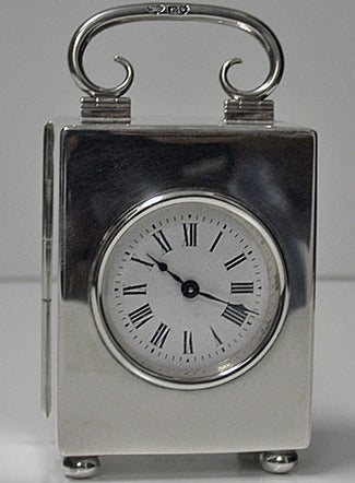 Silver carriage clock, London, 1913, by The Goldsmiths and Silversmiths Co. Ltd. The plain rectangular clock on four bun feet with hinged plain scroll design handle, solid hinged back cover with circular white enamel dial and black Roman numerals.