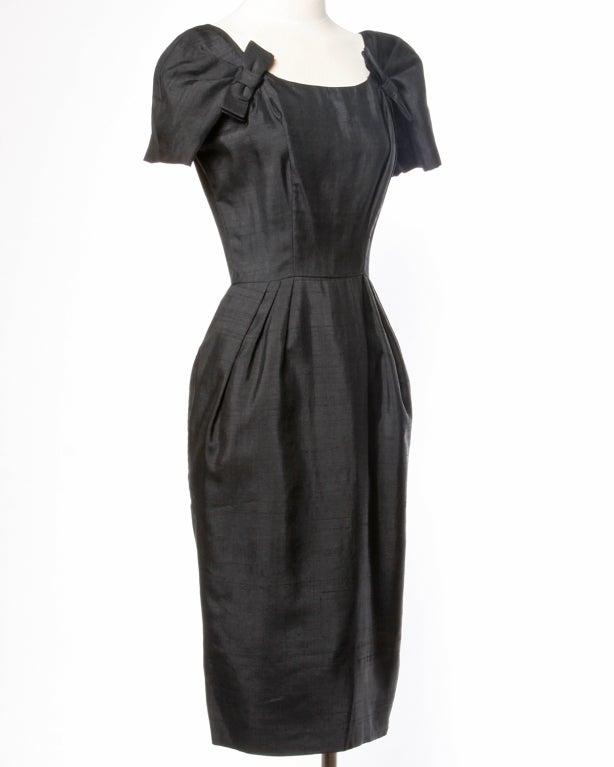 Simple but chic black raw silk wiggle dress by Suzy Perette. Nipped waist and short sleeves embellished with tiny bows. Fully lined with rear metal zip and hook closure. Classic 1950's silhouette! 

Bust: Up to 40