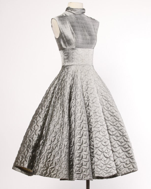 Incredible muted metallic silver vintage 1950s dress with a quilted skirt and classic fit and flare 50s silhouette. The dress features sleeveless sleeves, a high neckline, and glitter buttons up the back. It is unlined with rear zip and button