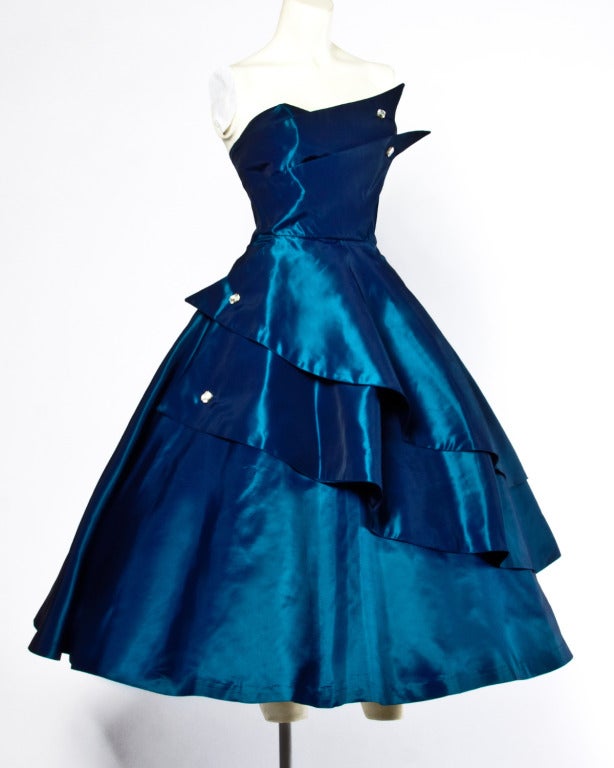 Stunning blue taffeta party dress from the 1950s featuring a unique asymmetric strapless cut with rhinestone embellishments. 

Details

Unlined
Back metal zip and hook closure
Circa: 1950s
Estimated Size: S
Colors: